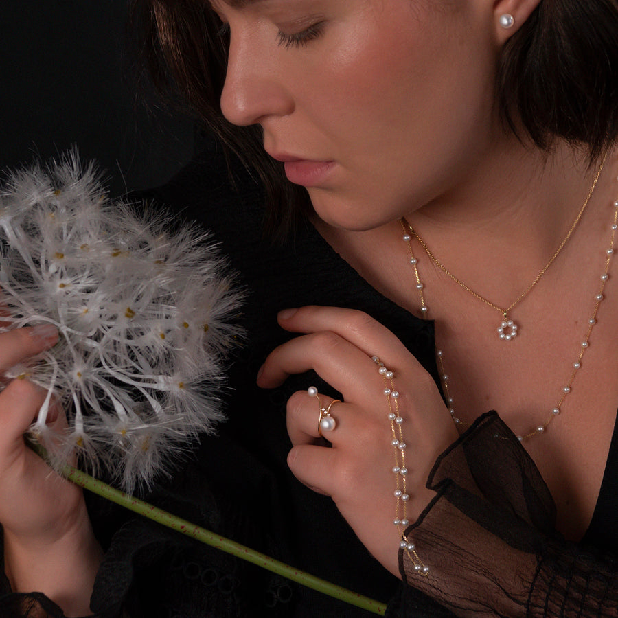 Nahaufnahme einer Frau, die zarten Schmuck von LA LARIS trägt. Sie hält eine große Pusteblume in der Hand und trägt den PEARL ORBIT Ring aus 750 Gold mit zwei Süßwasserperlen und drei Brillanten an der rechten Hand. Zudem sind eine zarte Halskette mit einem Perlenanhänger und Perlenohrstecker sichtbar.
